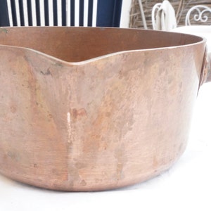 May include: A copper pot with a wooden handle. The pot has a rounded bottom and a wide, shallow opening. The copper has a warm, aged patina.