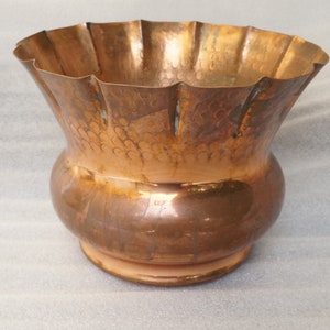 Puede incluir: Un jarrón de color cobre con un borde ancho y ondulado y una base bulbosa. El metal tiene una textura martillada en la parte superior y un acabado liso en la parte inferior. El jarrón mide aproximadamente 20 cm de alto.