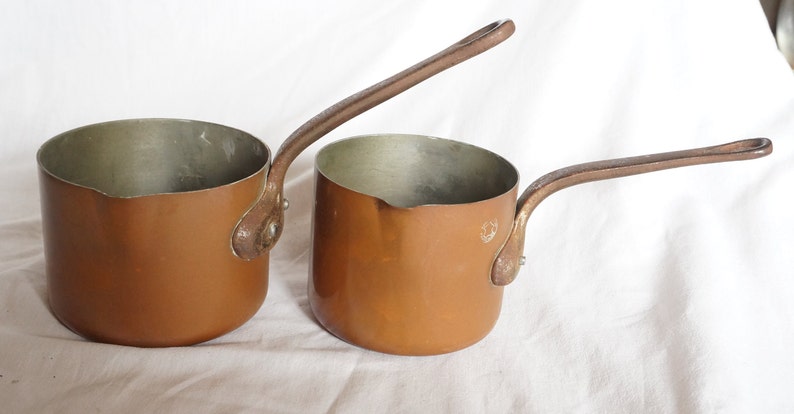 May include: Two copper sauce pans with long handles. The pans are different sizes and have a shiny, metallic finish.