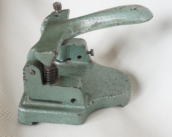 May include: A vintage green metal hole punch with a lever handle and a spring mechanism. The punch has a round hole for punching paper.