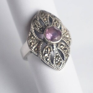 Puede incluir: Un anillo de plata con un delicado diseño de filigrana que presenta una piedra preciosa púrpura en el centro.