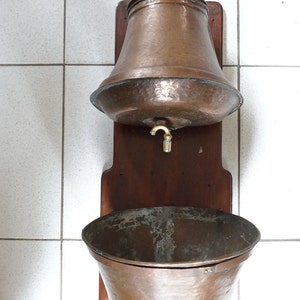 Puede incluir: Juego de lavabo antiguo de cobre. El juego incluye un recipiente de agua de cobre martillado con tapa y grifo, una tabla de montaje de madera y una pila de cobre. El cobre tiene una rica pátina envejecida. El juego está montado en una pared alicatada blanca.