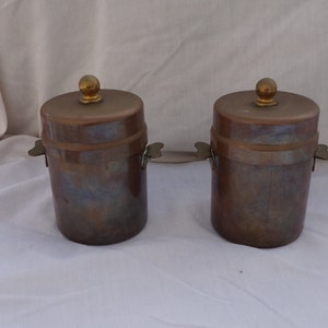 Può includere: Due barattoli di rame antichi con coperchi e manici in ottone. I barattoli hanno un aspetto usurato e invecchiato e sono un bellissimo esempio di utensili da cucina vintage.