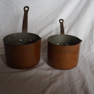 May include: Two copper sauce pans with handles. The pans are different sizes and have a shiny, metallic finish.