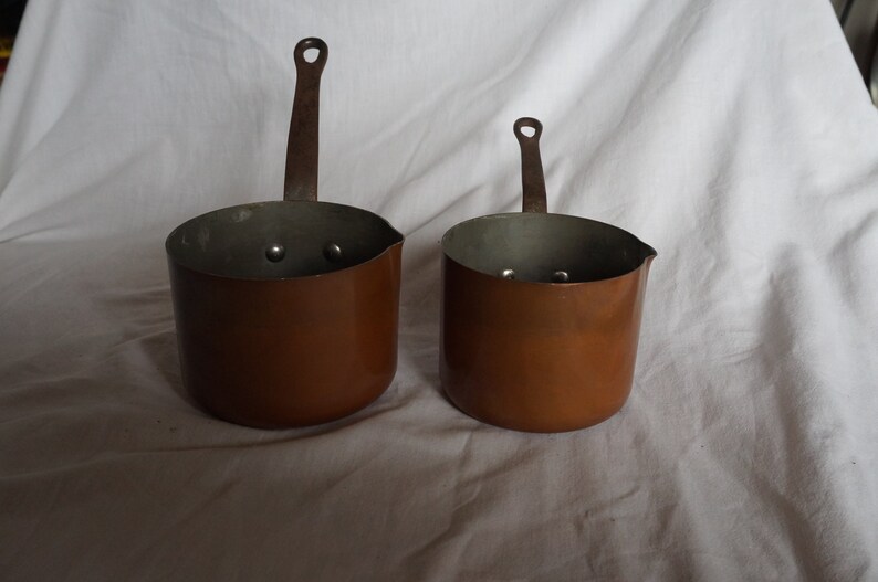 May include: Two copper sauce pans with handles. The pans are different sizes and have a shiny, metallic finish.