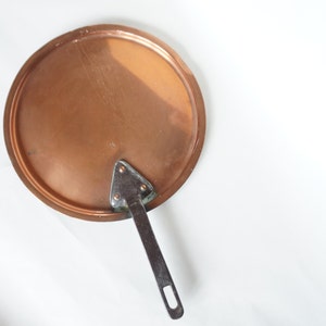 May include: A copper pot lid with a silver handle. The lid is round and has a smooth surface. The handle is attached to the lid with a triangular piece of metal.