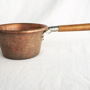 Puede incluir: Una cacerola de cobre con acabado martillado y mango de madera. La cacerola tiene dos asas de metal en los lados para levantarla.