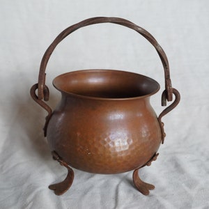 Puede incluir: Una olla de cobre martillada con un asa de hierro forjado y tres patas. La olla es de color marrón rústico y tiene un aspecto ligeramente envejecido.