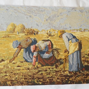 May include: A vintage needlepoint tapestry depicting three figures in a field harvesting wheat. The figures are wearing traditional clothing and are bending over to gather the wheat. The field is a golden yellow color and the sky is a light blue with clouds.