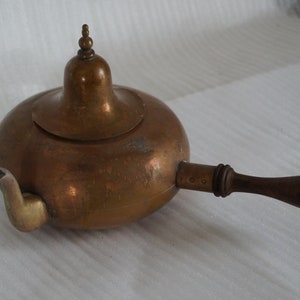 Op de afbeelding: Een koperen theepot met een houten handvat en een gewelfd deksel. De theepot heeft een tuit en een decoratief knopje op het deksel.