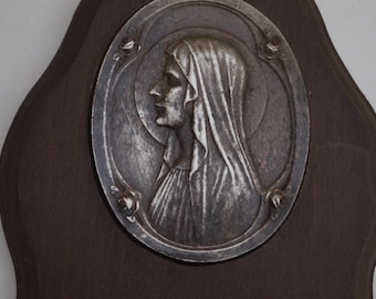 May include: A silver-toned metal plaque with a relief sculpture of a woman's head in profile. The plaque is mounted on a dark brown wooden background.
