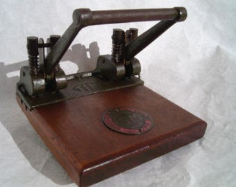 May include: Antique metal paper punch with a wooden base. The punch has a lever and two holes. The base has a circular logo with the text "The Acme Co."