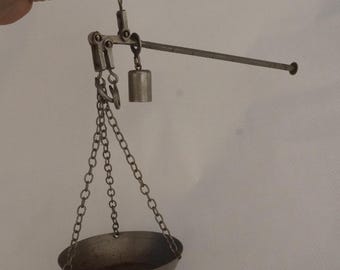 May include: A silver hanging scale with a bowl and a weight. The scale is made of metal and has a chain.