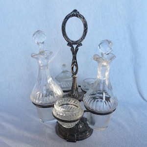 Könnte beinhalten: Ein versilbertes Gewürzset mit vier Glasbehältern. Das Set hat einen dekorativen Griff mit einer runden Öffnung und einen Sockel mit einem floralen Design.