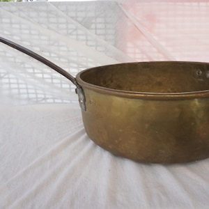 Peut inclure: Une casserole en laiton vintage avec une longue poignée en métal foncé. Le pot a une forme arrondie et un bord légèrement évasé. Le laiton a une teinte dorée chaude et la poignée est fixée avec un support métallique.