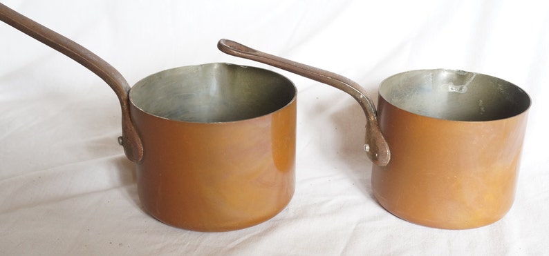 May include: Two copper saucepans with long handles. The saucepans are a warm copper color and have a shiny finish.