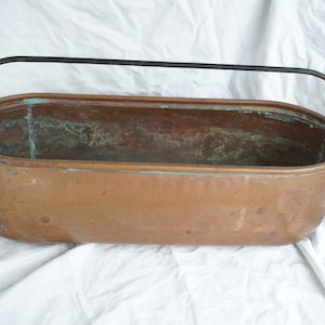 Può includere: Un vaso ovale in rame con un manico in metallo nero. Il vaso ha un aspetto rustico e invecchiato ed è perfetto per esporre piante o fiori.
