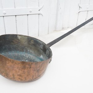 May include: A large copper pot with a long, black iron handle. The pot has a rounded bottom and a wide, shallow opening. The pot is old and has a patina of age.