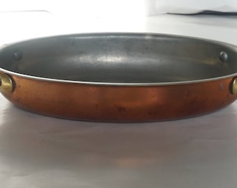 May include: A copper oval baking dish with brass handles. The dish is empty and has a shiny, metallic finish.