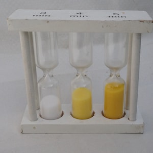 以下が含まれることがあります： 3分、4分、5分とラベル付けされた、黄色の砂が入った白い砂時計が3つあります