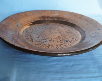 Pode incluir: Um prato de servir grande, redondo e de cobre com acabamento martelado e um padrão decorativo de pequenas folhas em relevo. O prato tem uma pátina marrom escura e uma borda ligeiramente elevada.