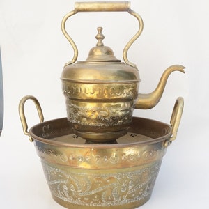May include: A vintage brass kettle with intricate floral designs sits atop a matching brass basin. The kettle has a spout and a handle, while the basin has two handles. Both pieces are ornately decorated with floral patterns.