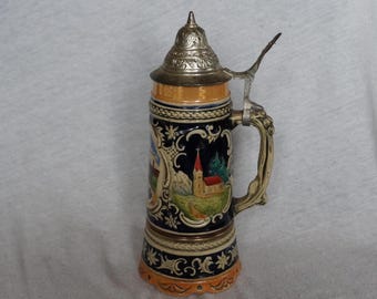 May include: A decorative beer stein with a silver-colored lid and handle. The ceramic body features a detailed landscape scene with a church, mountains, and trees, set against a dark blue background. The base and top rim have ornate patterns.