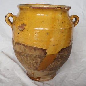 May include: An antique ceramic jar with a wide opening and two small handles. The jar has a two-tone design, with a golden-yellow upper section and a brown lower section. The surface shows signs of age and wear, adding to its vintage appeal.