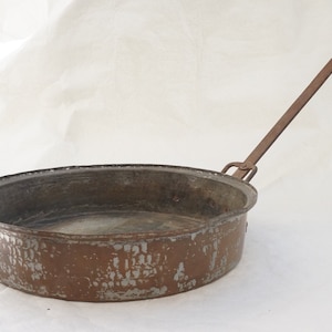 Peut inclure: Une grande poêle ronde en cuivre avec une longue poignée en fer courbée. La poêle a un aspect usé et vieilli avec une patine blanche et brune.