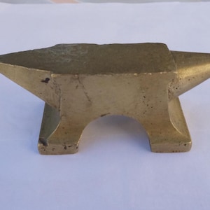 May include: A small, gold-colored metal anvil with a rounded top and a flat base. The anvil is about 4 inches long and 2 inches wide.