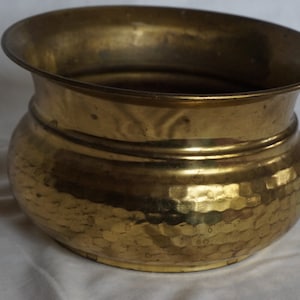 Può includere: Un vaso da fiori in ottone martellato con una base larga e arrotondata e un bordo svasato. Il vaso ha una finitura martellata testurizzata ed è di colore dorato caldo.