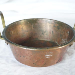 Puede incluir: Un pequeño cuenco de cobre antiguo con dos asas de latón. El cuenco tiene forma redondeada y un aspecto ligeramente deslustrado, con desgaste y pátina visibles. Las asas son arqueadas y están unidas a los lados del cuenco.