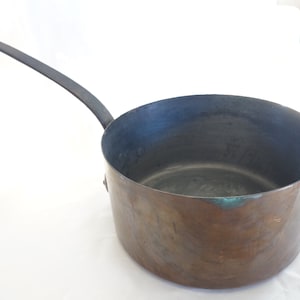 Peut inclure: Une casserole en cuivre avec une poignée en métal noir. La casserole a un aspect usé et vieilli et est légèrement ternie.