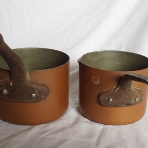 May include: Two small copper pots with brown handles. The pots have a light brown interior and a darker brown exterior.