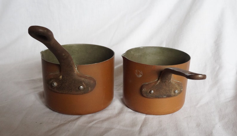 May include: Two small copper pots with brown handles. The pots have a light brown interior and a darker brown exterior.