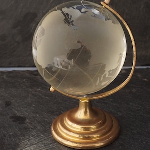 Puede incluir: Un pequeño globo de vidrio con un soporte de metal dorado. El globo es transparente y muestra los continentes en un diseño esmerilado.