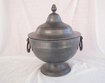 May include: A pewter-toned lidded serving bowl with a pedestal base and two ring handles. The lid has a small knob on top. The bowl is likely made of metal and sits on a white surface.