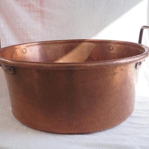 Puede incluir: Una olla grande de cobre martillado con dos asas resistentes. La olla tiene una parte superior ancha y abierta y una base redondeada. El cobre tiene un tono marrón rojizo cálido, y la superficie muestra signos de edad y uso. Esta olla vintage es adecuada para cocinar o exhibir.