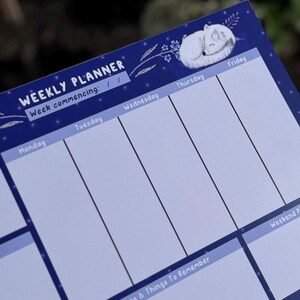 May include: A dark blue weekly planner featuring a sleeping white fox illustration. The planner is divided into sections for each day of the week, with the text "WEEKLY PLANNER" and "Week commencing:" at the top.