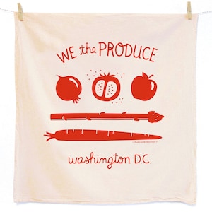 May include: A cream-coloured kitchen towel with red text and illustrations of produce. The text reads "WE the PRODUCE washington D.C." and features a pomegranate, tomato, apple, asparagus, and carrot.
