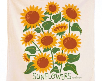 Op de afbeelding: Een crèmekleurige theedoek met een ontwerp van gele zonnebloemen met bruine centra en groene bladeren. De tekst "SUNFLOWERS" is in zwart gedrukt aan de onderkant van de theedoek. De tekst "The Neighborgoods" is in zwart gedrukt in de rechterbenedenhoek van de theedoek.