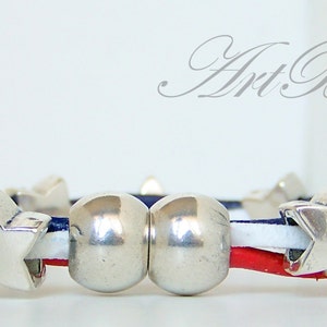 May include: A red, white, and blue leather bracelet with silver star and bead charms. The bracelet is made with three strands of leather, each a different color. The bracelet has two silver beads and several silver star charms.