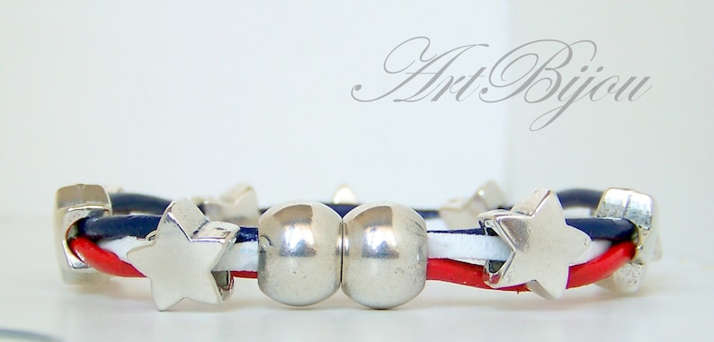 May include: A red, white, and blue leather bracelet with silver star and bead charms. The bracelet is made with three strands of leather, each a different color. The bracelet has two silver beads and several silver star charms.