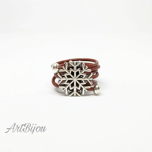 Peut inclure: Une bague en cuir marron avec un charme en forme de flocon de neige argenté. La bague est réglable et peut être portée à n'importe quel doigt.