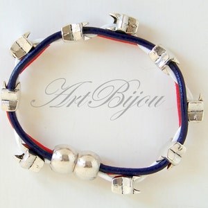 May include: A blue, white, and red leather bracelet with silver metal charms. The charms are shaped like arrows and there is a double silver bead in the center.