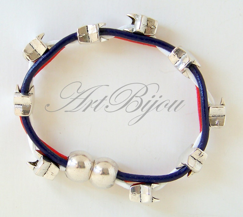 May include: A blue, white, and red leather bracelet with silver metal charms. The charms are shaped like arrows and there is a double silver bead in the center.