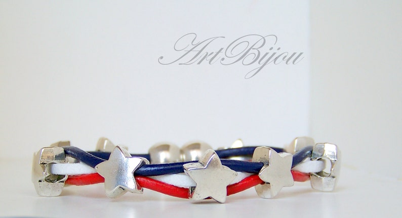 May include: A red, white, and blue leather bracelet with silver star charms. The bracelet is woven with three strands of leather, each a different color. The stars are evenly spaced along the bracelet.