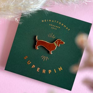 Könnte beinhalten: Ein Dackel-Anstecknadel mit braunem Körper und grünem Kragen auf einer dunkelgrünen Karte. Die Karte trägt goldfarbene Schriftzüge: "HEIMATFORMAT MÜNCHEN 2014" und "SUPERPIN". Der Hintergrund ist rosa.
