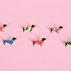 Könnte beinhalten: Fünf Dackel-Pins in verschiedenen Farben und Mustern. Die Pins sind alle schwarz mit goldenen Umrissen und haben verschiedene farbige Pullover. Die Pullover sind rot, blau, grün, gelb und rot-weiß gestreift.