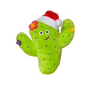 Op de afbeelding: Een groene cactus knuffel met een lachend gezicht, versierd met kleine bloemen en een rood-witte kerstmuts. De cactus heeft twee armen en is ontworpen om eruit te zien als een vriendelijk karakter.